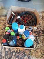 Two cardboard boxes containing battery cables, automotive and household cleaning products, motor oil and transmission fluid, arranged on carpet.