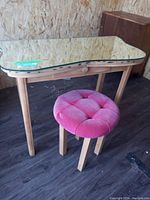 Wooden vanity table topped with a curved glass sheet, showcasing a pink round cushioned stool beneath it.