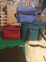 Three insulated cooler bags stacked: red, blue on top, and large teal Thermos bag at bottom on a table.