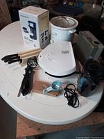 Wide view of all kitchen appliances on white table including coffee maker box, crockpot, electric heater, George Foreman grill, knives with wooden block, hand mixer, kettle, and egg separator.