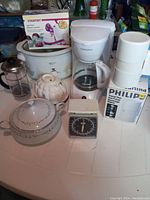 Photo showing white Crockpot with floral design, 12 cup drip coffee maker, coffee press, kitchen scale, mould and serving dish, and new apple peeler in box.