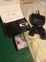 Open box with Seiki Cambre media streaming box, black binoculars, and retail box for wireless earbuds on a green surface.