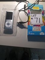 Photo of the silver MP4 player in clear packaging beside the colorful ZYZY brand box and USB charging cable.