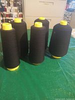 Five large cones of black serging or sewing thread with yellow spool bases arranged on a green surface.