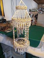 Full view of the shell plant hanger hanging over a table, showing its length, conical top, and shell strands hanging down with circular base.