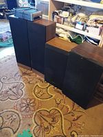 Five wooden cabinet speakers with black grills arranged side by side on a patterned rug, basement storage background