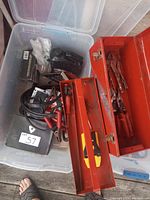 Red metal toolbox with lid open, showing hand tools inside including locking pliers and screwdrivers. Plastic storage tote visible underneath containing booster cables and other items.