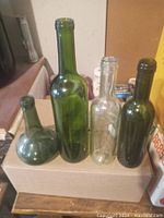 Four empty wine bottles of assorted sizes and shapes placed on a cardboard box, showing variety in bottle design and color including green and clear glass.