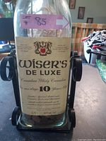 Front view of Wiser's De Luxe whisky jug showing label and black metal swinging stand holding it.