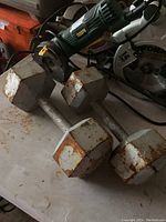 Photo of two 25 pound hexagonal dumbbell weights connected by a metal handle on a workshop surface, showing signs of rust.
