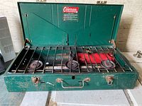 Photo showing green Coleman stove with lid open exposing three burners and black metal grate. The stove appears used with visible wear marks.