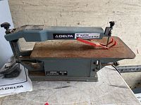 Side view of the Delta 15-inch scroll saw showing the metal work surface, blue body, and blade with guard.