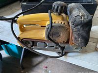 Side view of yellow DeWalt belt sander showing power cord and tension knob, placed on white surface.
