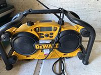 Front view of yellow and black DeWalt radio with dual speakers, control buttons, and attached power cord inside a protective black metal frame.