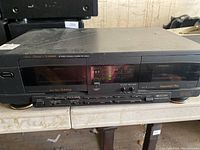 Front view of black Fisher stereo double cassette deck with Synchro Dubbing and Sequential Play written on it. Unit appears powered on showing some lights on the display.
