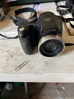 Black Fujifilm FinePix S700 digital camera with attached strap shown resting on a white surface, close-up view of camera front and top.