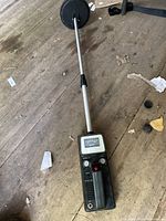 Full view of the metal detector showing the black body with controls, analog meter, and circular search coil on a long metal shaft laying on a wooden floor.