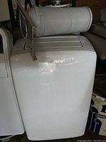 Front and top view of white NOMA portable standing air conditioner unit with attached vent hose and window vent panel, showing overall condition and completeness of the item.