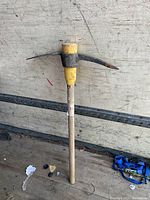 Front view of pickaxe showing yellow and black metal head attached to wooden handle with some wear and chipping paint.