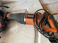 Ridgid reciprocating saw sitting on a wooden surface with visible power cord wrapped around the handle area, orange and black color scheme.