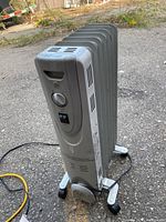 Photo of gray oil-filled radiator space heater standing on concrete ground with visible power cord and caster wheels.
