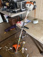 Full view of Stihl FS 38 gas powered string trimmer standing upright on a concrete floor next to a table with various items on it