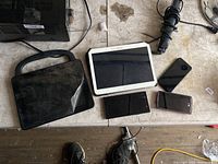 Photo shows two tablets and three smartphones on workbench. Tablets: Samsung white tablet and black tablet with cover. Phones: black folding phone, black smartphone, pink metallic smartphone.