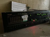 Photo of the Teac EQ-305L stereo graphic equalizer powered on, showing LED indicators lit across frequency sliders and control buttons on the front panel.
