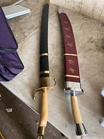 Two Japanese swords laid on a table with their sheaths. Black sheath with gold and brass accents, and a maroon sheath with gold Japanese characters with silver ends. Light-colored handles on both swords.