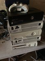 Photo showing five Xbox 360 consoles stacked with a white wireless controller on top, various colors visible including black, white and gray.