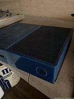 Front and left side view of blue Xbox One console with visible round power button and vent slots.