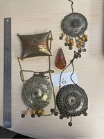 Photo showing three circular metal pendant necklaces with decorative embossing and hanging beads, small metal case with chain, and amber teardrop pendant.