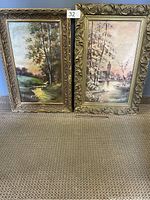 Two original oil paintings on canvas with wood frames placed on carpet; one painting shows a green landscape with trees and sunlight, the other depicts a water scene with trees and a distant tower under a pastel sky.