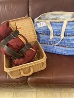 Photo of wicker picnic basket open with two rolled picnic blankets inside next to blue insulated beach bag with sailboat pattern on brown leather sofa.