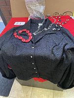 Photo of a black textured ladies blouse with pearl-like buttons and assorted costume jewelry including a chunky red bead bracelet and several necklaces with silver chain, black, white, and pearl-like beads.