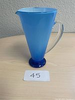 Single image showing a blue retro glass pitcher on a flat surface against a neutral background with a lot number card in front.