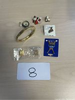 Photo showing a gold-finish bangle bracelet with screw detail, a pearl and blue enamel swirl pin, a Canadian flag and dancing couple enamel pin, a white enamel flower pin, a vintage-style clothing-themed pin on card, a blue enamel key shaped pin on card, and a pair of small gold-tone clip-on earrings still in plastic.