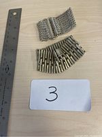 Two metal elasticized bracelets placed near a ruler for size reference on a wooden surface. One bracelet has a wide mesh chain design, the other has horizontal bar links.