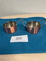 Two small copper pots with stainless steel interiors positioned on blue cloth, showing exterior and handles