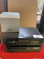 Front and top view of two black Sony electronic VHS/cassette players stacked with a white Dell computer tower on top in natural light near window.