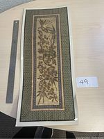 Photo of the large Chinese embroidered wall hanging in original packaging placed on a light wood surface, with a ruler for size comparison and a paper label marked 49.