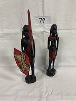 Two hand carved Kenyan figurines shown side by side, a warrior with spear and shield and a woman figurine.