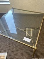 Glass top coffee table with brass frame, shows corner finials and circular structural support underneath.
