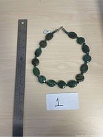 Photo of a green stone necklace placed next to a ruler for size reference on a light wooden surface.