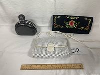 Three ladies evening bags: small black rhinestone clutch, black velvet floral embroidered clutch, white woven handbag with cream tab; ruler included for scale; bags show animal damage