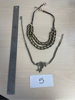 Photo shows two necklaces laid flat on a light surface alongside a ruler for size reference. One necklace has thick metal chain links combined with leather bands, the other is an aged tribal metal chain with a decorative pendant and tassels.