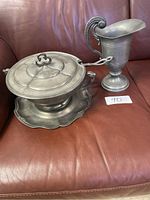 Photo showing the pewter soup tureen with lid, ladle resting on tray, and pitcher together on a couch.