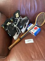 Pair of black and light blue girls hockey skates by Mission, size 6 (37), with laces, seated on a red couch next to a wooden badminton racket with synthetic strings and leather handle and a box of poker chips with playing cards.