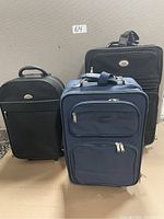 Three carry on suitcases: two black and one navy blue. Each has wheels, handles and exterior zipper compartments.