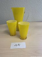 Photo showing yellow glass pitcher and two matching glasses on a gray surface in front of a gray wall with a number card '44' in front.